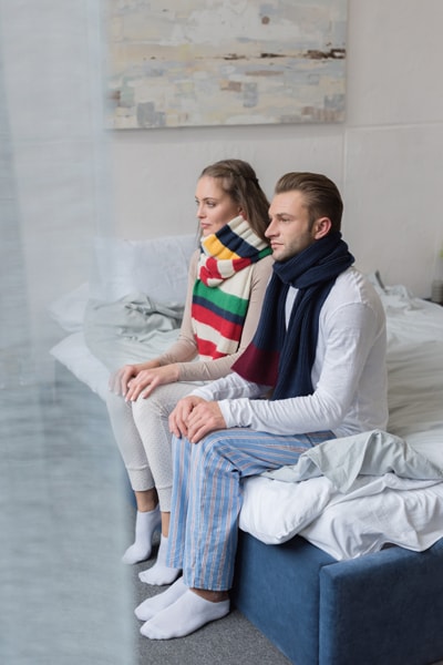 Sick couple sitting in pajamas on a bed with scarfs over necks.